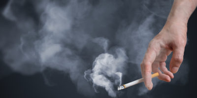 Smoker is holding smoking cigarette in hand. A lot of smoke around.